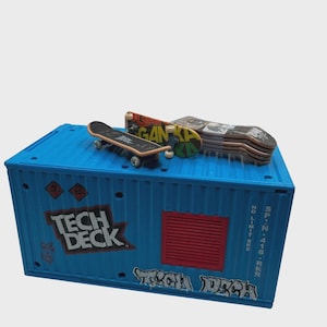 May include: A blue Tech Deck container with several miniature skateboards on top. The container has the words "TECH DECK" in white and red graffiti-style lettering. The skateboards have various colorful designs.