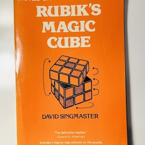 May include: An orange book cover with the title "Notes on Rubik's Magic Cube" and the author's name "David Singmaster". The cover features a black and white illustration of a Rubik's Cube with arrows indicating how to turn the sides.
