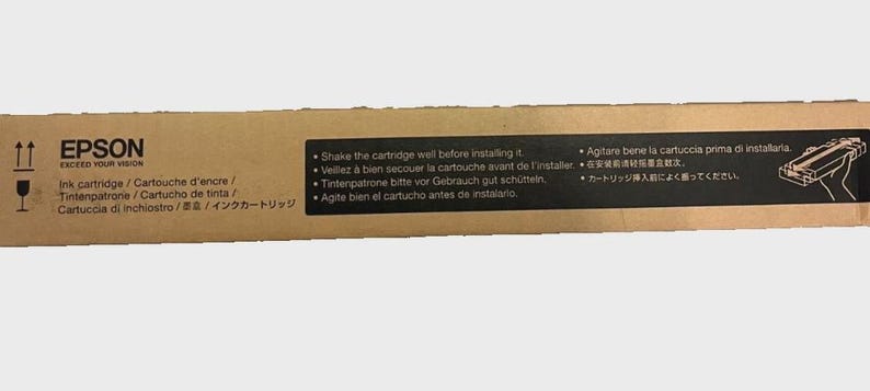 May include: A brown cardboard box with the Epson logo and the words "EXCEED YOUR VISION." The box contains an ink cartridge and instructions in multiple languages, including English, French, German, Spanish, Italian, and Japanese.