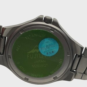 May include: A close-up of the back of a silver wristwatch. The watch face is green with the text "TITANIUM", "FUJITIME JAPAN", and "M2505QT". A blue sticker with the text "FUJITIME F.T.R" is also visible. The watch is water resistant.