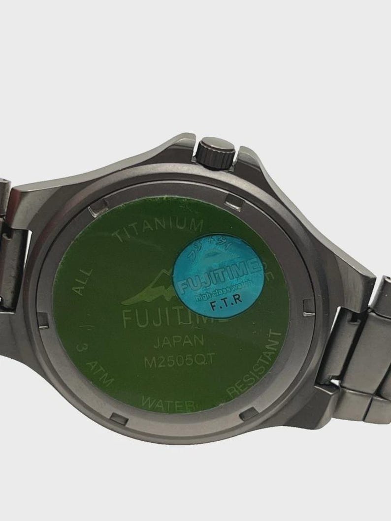 May include: A close-up of the back of a silver wristwatch. The watch face is green with the text "TITANIUM", "FUJITIME JAPAN", and "M2505QT". A blue sticker with the text "FUJITIME F.T.R" is also visible. The watch is water resistant.