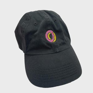 May include: Black baseball cap with a curved brim. The cap features an embroidered donut logo with a pink frosting and yellow center. The hat is made of a durable fabric and has a classic, casual style.