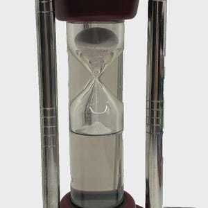 May include: An hourglass with a dark red wooden frame and silver metal supports. The glass chamber contains white sand and is designed to measure the passage of time. The hourglass is a decorative item.