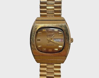 May include: A vintage gold-toned wristwatch with a rectangular face and a matching band. The watch face displays the day and date, with the brand name "CLINTO" visible. The watch has a self-winding mechanism.