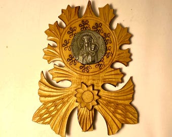 May include: A wooden religious icon featuring a silver medallion with a depiction of the Virgin Mary and child. The icon has a carved floral design with a sun-like element at the bottom. The wood has a light brown color.