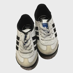 May include: A pair of white and black Adidas sneakers for toddlers. The shoes have black stripes on the sides, black accents, and white laces. The soles are beige and black.