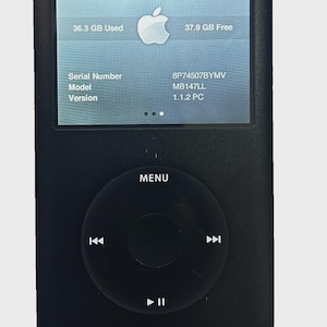 May include: Black iPod Classic with a white Apple logo on the screen. The screen displays storage information: 36.3 GB used, 37.9 GB free. The device has a click wheel and a menu button. A white charging cable is connected.