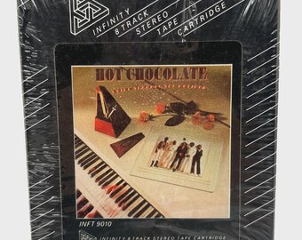 May include: A sealed black Infinity 8-track stereo tape cartridge featuring the album "Hot Chocolate." The album art shows a piano, a group of people, and a musical instrument. The cartridge is wrapped in clear plastic.