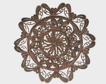 May include: A circular copper-toned metal filigree ornament featuring an intricate openwork design. The piece displays a central radial pattern surrounded by symmetrical floral and leaf-like motifs, creating a delicate, lace-like appearance.