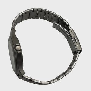 May include: A dark grey wristwatch with a metal strap. The watch face is round and dark, with a small button on the side. The strap is made of small, rectangular links. The watch is shown from a side angle.