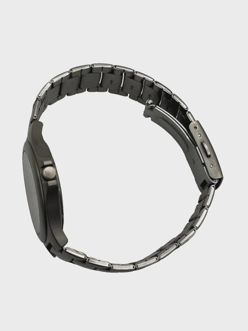 May include: A dark grey wristwatch with a metal strap. The watch face is round and dark, with a small button on the side. The strap is made of small, rectangular links. The watch is shown from a side angle.