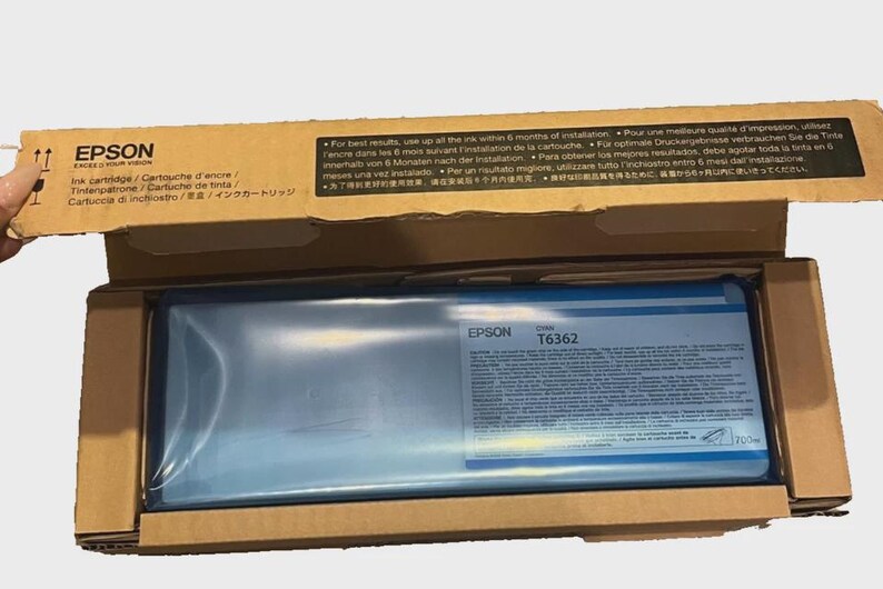 May include: Epson T6362 ink cartridge in a cardboard box. The box is open, revealing the ink cartridge inside a blue plastic wrap. The Epson logo and product information are visible on the cartridge.