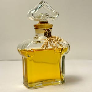 May include: A clear glass perfume bottle with a heart-shaped stopper. The bottle contains a golden liquid and is sealed with a gold-colored cap and a decorative tassel. The bottle has a vintage aesthetic.