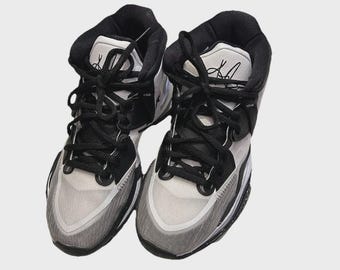 May include: Pair of high-top sneakers in black, white, and gray. The shoes feature black laces, a padded black collar, and a white and gray toe box. The tongue has a signature in black.