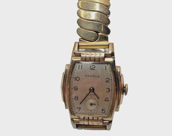May include: Vintage gold-toned Benrus watch with a rectangular face and a stretch band. The watch face has black numerals and hands, with a small seconds dial. The band is made of curved, interlocking metal links.