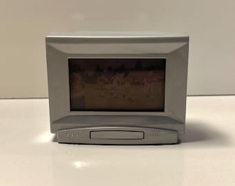 May include: A small, silver-colored digital photo frame. The frame displays a picture with people in it. The frame has a rectangular screen and a small control panel at the bottom. The frame is sitting on a white surface.