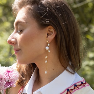 Puede incluir: Una mujer lleva una camisa blanca con un patrón bordado de colores en el cuello. Lleva pendientes largos y colgantes con perlas blancas y una cadena dorada.