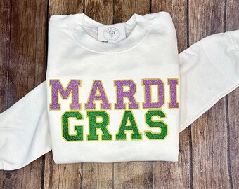 May include: White sweatshirt with the words "MARDI GRAS" in purple and green glitter lettering. The long-sleeved top is folded on a wooden surface. A small tag is visible near the collar.