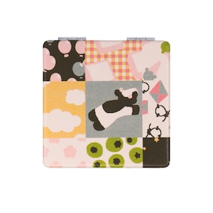 May include: A small, square, pink and black compact mirror with a patchwork design. The design features a black and white cow, pink and white polka dots, a pink and white checkered pattern, and green kiwi fruit.