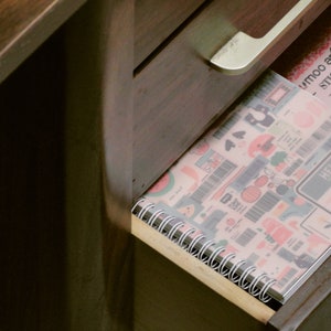 May include: A partially opened wooden drawer reveals a spiral-bound notebook and a sticker book. The notebook features a colorful pattern with various illustrations, while the sticker book has a pink cover with a floral design. The text "HELLO" is visible on the notebook.
