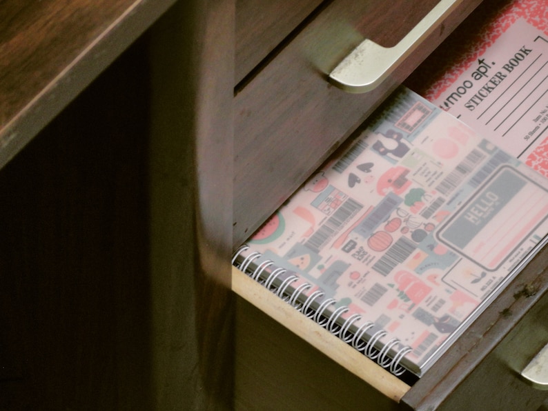 May include: A partially opened wooden drawer reveals a spiral-bound notebook and a sticker book. The notebook features a colorful pattern with various illustrations, while the sticker book has a pink cover with a floral design. The text "HELLO" is visible on the notebook.