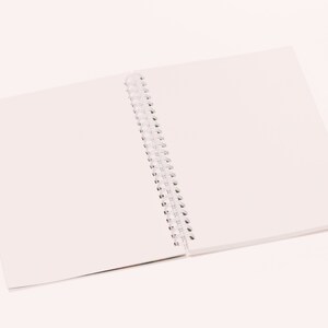 May include: An open, spiral-bound notebook with blank white pages. The notebook is lying flat on a white surface, with the pages spread open to reveal the clean, unused paper. The spiral binding is visible along the spine.