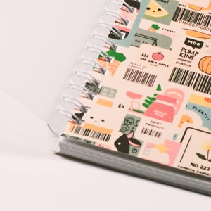 May include: A spiral-bound notebook with colorful, illustrated pages. The pages feature various food-themed graphics, including carrots, pumpkins, and dairy products, along with text such as "Gala Apple" and "Organic Produce."