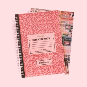 May include: A spiral-bound sticker book with a pink cover featuring a speckled pattern. The cover has a label that reads "STICKER BOOK" and "50 sheets". A second sheet with colorful graphics is visible behind the book.