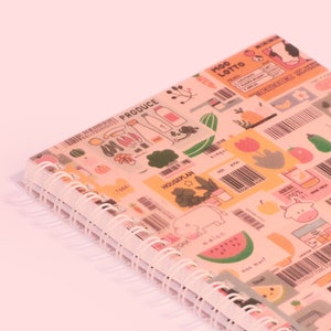May include: A spiral-bound notebook with a colorful cover featuring illustrations of produce, barcodes, and the text "MOO LOTTO". The notebook has a white spiral binding and is set against a light pink background.