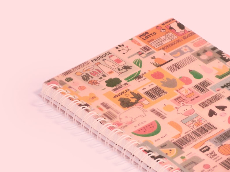 May include: A spiral-bound notebook with a colorful cover featuring illustrations of produce, barcodes, and the text "MOO LOTTO". The notebook has a white spiral binding and is set against a light pink background.
