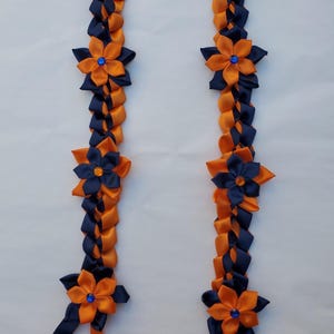 May include: A braided ribbon lei in navy blue and orange. The lei features flower accents with blue gem centers. The ribbon is woven in a repeating pattern. The lei is displayed against a white background.