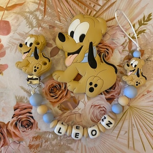 Peut inclure: Un hochet pour bébé en silicone bleu et beige avec un clip, représentant un petit et un grand chien de dessin animé brun et jaune. Le hochet est doté d'une chaîne de perles avec le nom "LEON" écrit en lettres blanches sur des blocs beiges.