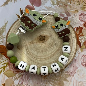 Peut inclure: Un hochet pour bébé en silicone vert et marron avec le nom "ZATHAN" écrit en lettres blanches. Le hochet présente deux petites figurines en silicone vert d'un personnage fictif populaire.