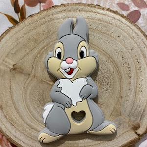Peut inclure: Une sucette en silicone grise et beige en forme de lapin de dessin animé avec une découpe en forme de cœur. Le lapin a un cœur blanc sur la poitrine et une petite découpe en forme de cœur sur le ventre.