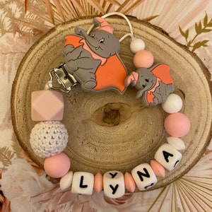 Peut inclure: Un hochet pour bébé en silicone rose et blanc avec une boule en crochet et un clip. Le hochet porte le nom "LYNA" en lettres noires sur des perles blanches. Le hochet est également décoré de deux breloques en forme d'éléphant en silicone.