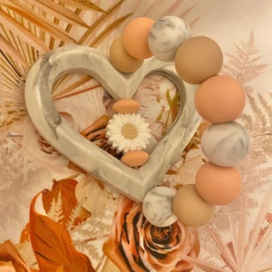 May include: A white heart-shaped silicone teether with a white daisy charm and a bracelet of beige and peach silicone beads with a marble pattern.
