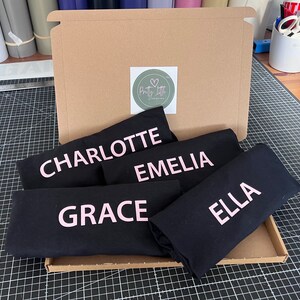 May include: A cardboard box filled with five black t-shirts, each with a different name printed in pink lettering. The names are Charlotte, Emelia, Grace, Ella, and Pretty Little Printing.