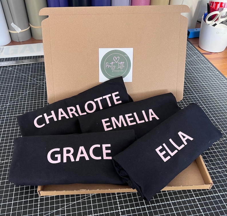 May include: A cardboard box filled with five black t-shirts, each with a different name printed in pink lettering. The names are Charlotte, Emelia, Grace, Ella, and Pretty Little Printing.