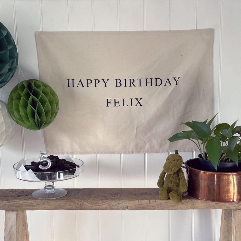 K&ouml;nnte beinhalten: Ein beigefarbenes Banner mit dem Text "Happy Birthday Felix" in schwarzen Buchstaben. Eine Holzbank mit einem gr&uuml;nen Stoffdinosaurier und einem Kupfertopf mit einer gr&uuml;nen Pflanze. Ein gl&auml;serner Kuchenst&auml;nder mit einem Schokoladenkuchen und einer silbernen Kerze mit der Nummer 5.