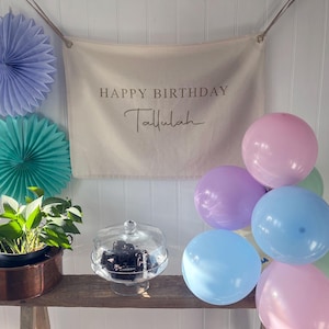 K&ouml;nnte beinhalten: Ein wei&szlig;es Banner mit dem Text "Happy Birthday Tallulah" h&auml;ngt an einer wei&szlig;en Wand. Eine Gruppe von pastellfarbenen Ballons in Rosa, Lavendel, Hellblau und Gr&uuml;n sind in einem Halbkreis unter dem Banner angeordnet. Eine kleine Topfpflanze mit gr&uuml;nen Bl&auml;ttern steht auf einem Holztisch vor dem Banner. Ein gl&auml;serner Kuchenst&auml;nder mit einem schwarzen Kuchen steht auf dem Tisch.