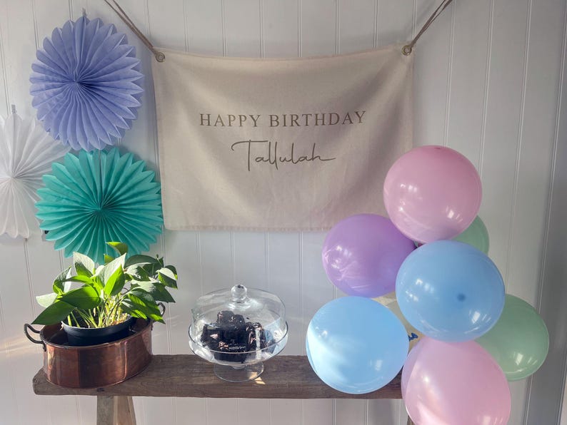 K&ouml;nnte beinhalten: Ein wei&szlig;es Banner mit dem Text "Happy Birthday Tallulah" h&auml;ngt an einer wei&szlig;en Wand. Eine Gruppe von pastellfarbenen Ballons in Rosa, Lavendel, Hellblau und Gr&uuml;n sind in einem Halbkreis unter dem Banner angeordnet. Eine kleine Topfpflanze mit gr&uuml;nen Bl&auml;ttern steht auf einem Holztisch vor dem Banner. Ein gl&auml;serner Kuchenst&auml;nder mit einem schwarzen Kuchen steht auf dem Tisch.
