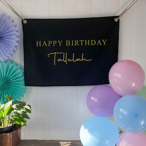 K&ouml;nnte beinhalten: Ein schwarzes Banner mit dem Text "Happy Birthday Tallulah" in goldenen Buchstaben. Das Banner h&auml;ngt an einer wei&szlig;en Wand mit blauen und gr&uuml;nen Papierf&auml;chern und pastellfarbenen Luftballons.