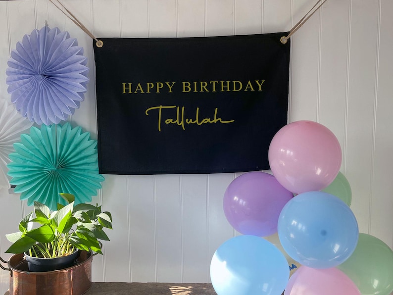 K&ouml;nnte beinhalten: Ein schwarzes Banner mit dem Text "Happy Birthday Tallulah" in goldenen Buchstaben. Das Banner h&auml;ngt an einer wei&szlig;en Wand mit blauen und gr&uuml;nen Papierf&auml;chern und pastellfarbenen Luftballons.