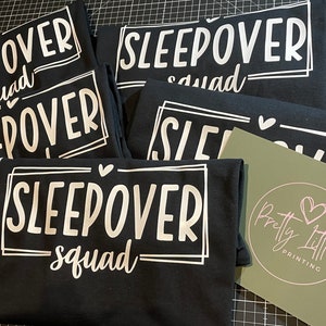 May include: Five black t-shirts with white text that reads "Sleepover Squad" in a fun, handwritten font. The shirts are stacked on top of each other, and a small green card with a pink heart logo is visible in the bottom right corner.