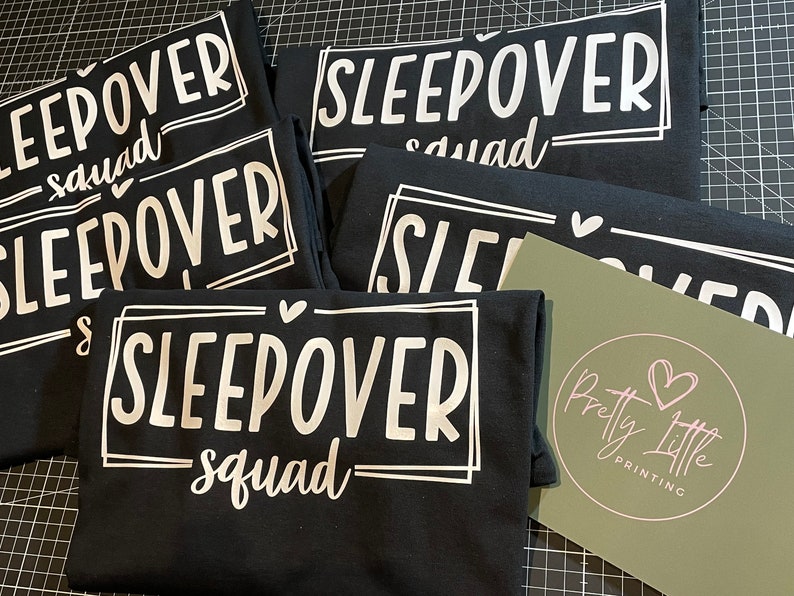 May include: Five black t-shirts with white text that reads "Sleepover Squad" in a fun, handwritten font. The shirts are stacked on top of each other, and a small green card with a pink heart logo is visible in the bottom right corner.