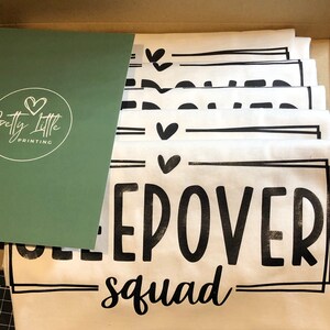May include: A stack of white t-shirts with black text that reads "Sleepover Squad" and a small heart graphic. The shirts are folded and stacked in a box.