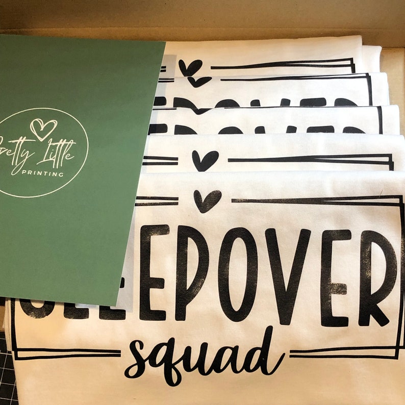 May include: A stack of white t-shirts with black text that reads "Sleepover Squad" and a small heart graphic. The shirts are folded and stacked in a box.