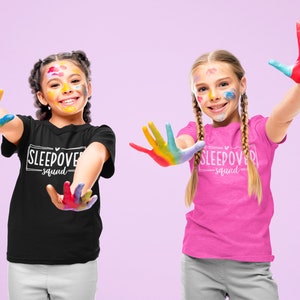 May include: Two children wearing black and pink t-shirts with the text "Sleepover Squad" printed on them. They are smiling and have colorful paint on their hands and faces.