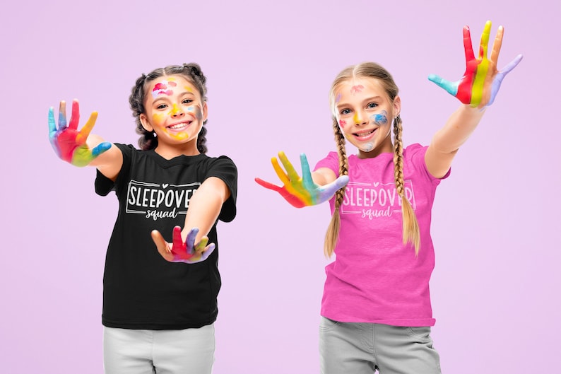 May include: Two children wearing black and pink t-shirts with the text "Sleepover Squad" printed on them. They are smiling and have colorful paint on their hands and faces.