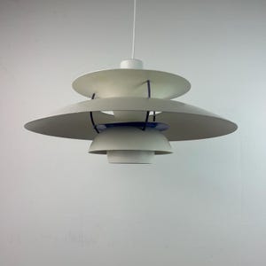 May include: A multi-tiered pendant light fixture featuring three stacked, circular, off-white metal shades. The shades are connected by thin, dark blue vertical supports, with a small blue accent ring visible between the lower two tiers.